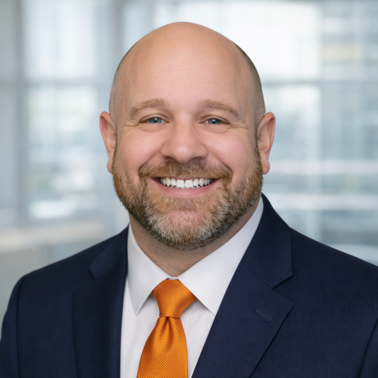 professional headshot of the owner and CEO of Abstract Health Solutions, shown smiling and wearing a navy suit, white shirt, and orange tie against a softly blurred office background.